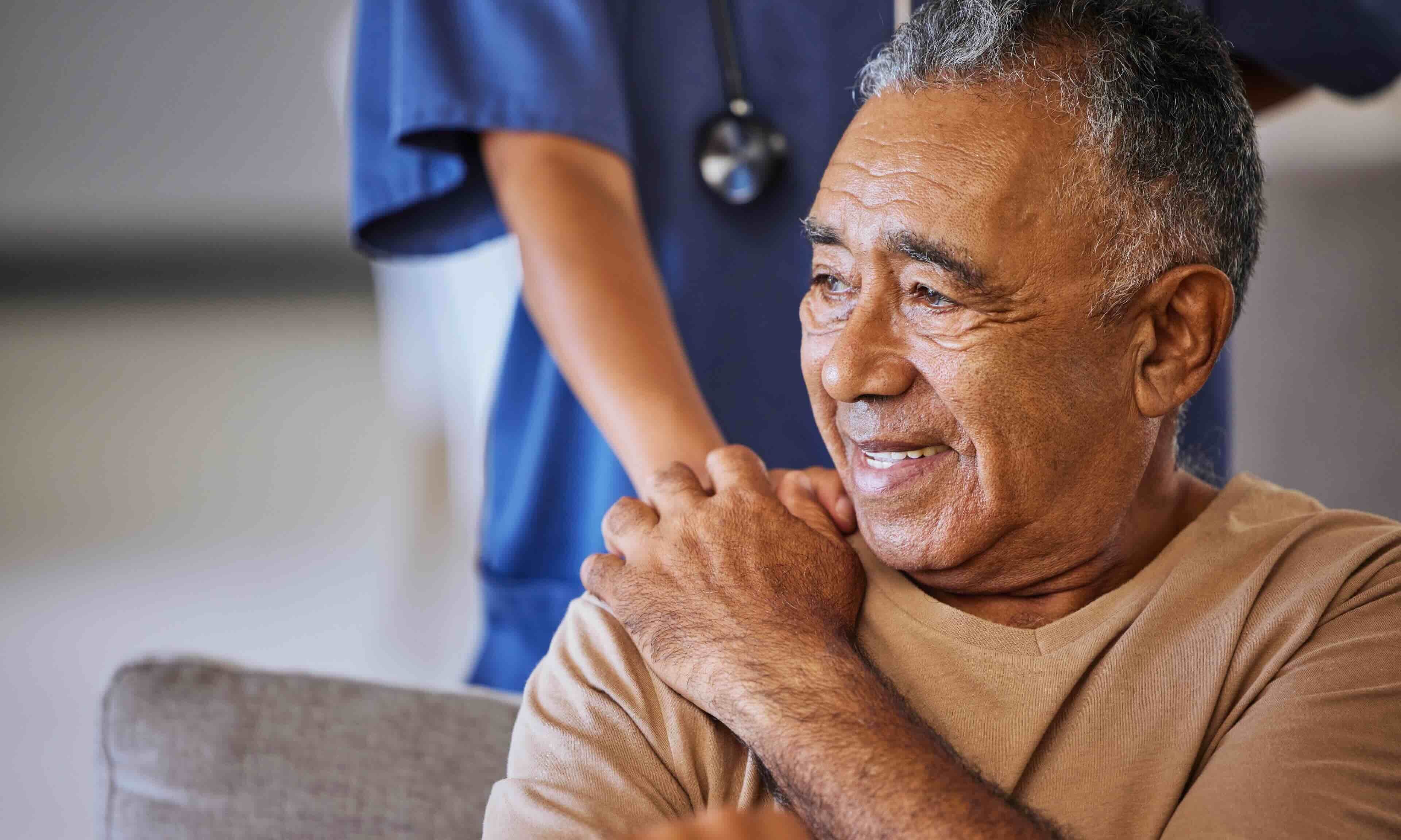 An elderly man smiling warmly while seated, with a healthcare worker in blue scrubs standing behind him and gently placing their hands on his shoulders.