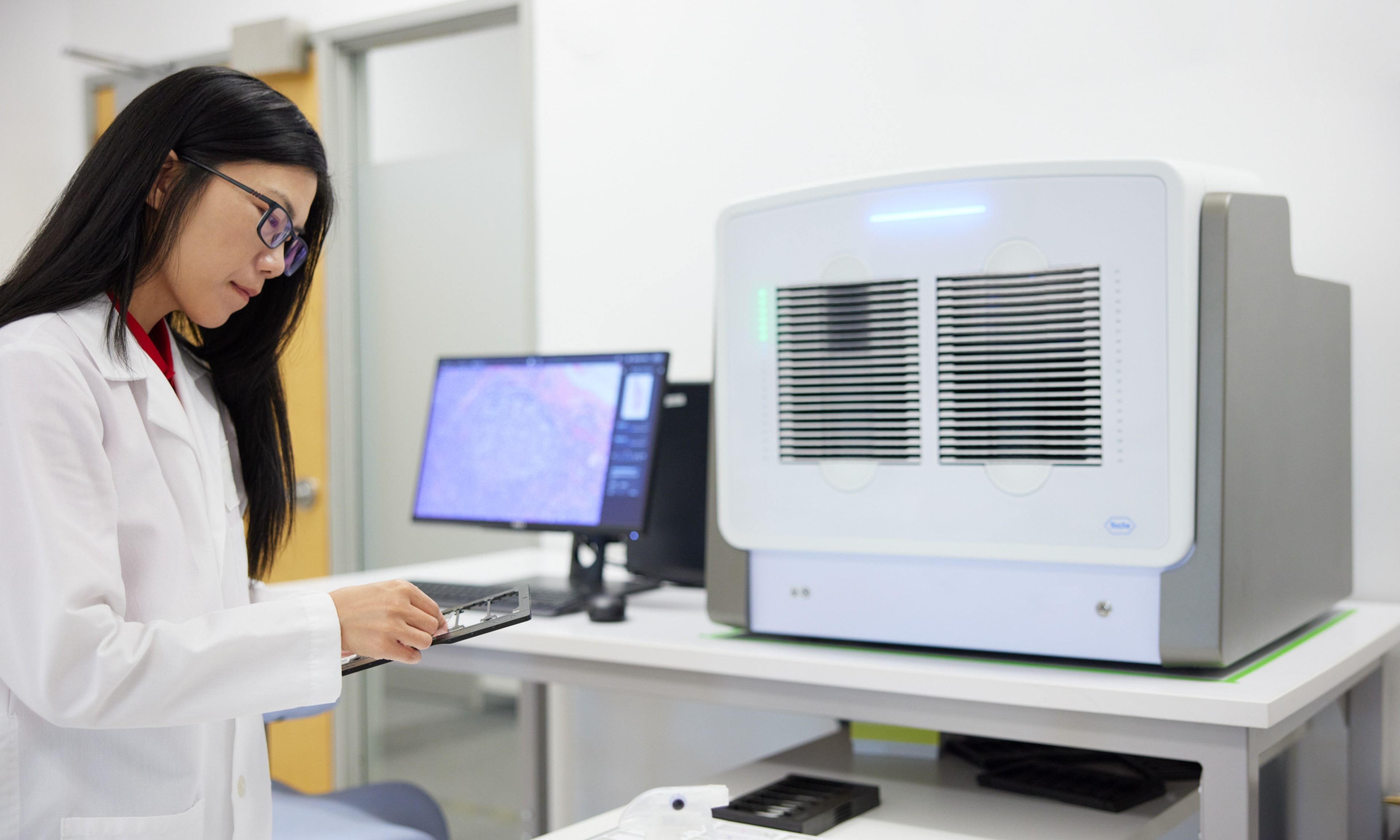 A laboratory technician working with a slide, wearing a white coat and glasses. Behind her, a computer screen displays microscopic images, and advanced laboratory equipment is visible.