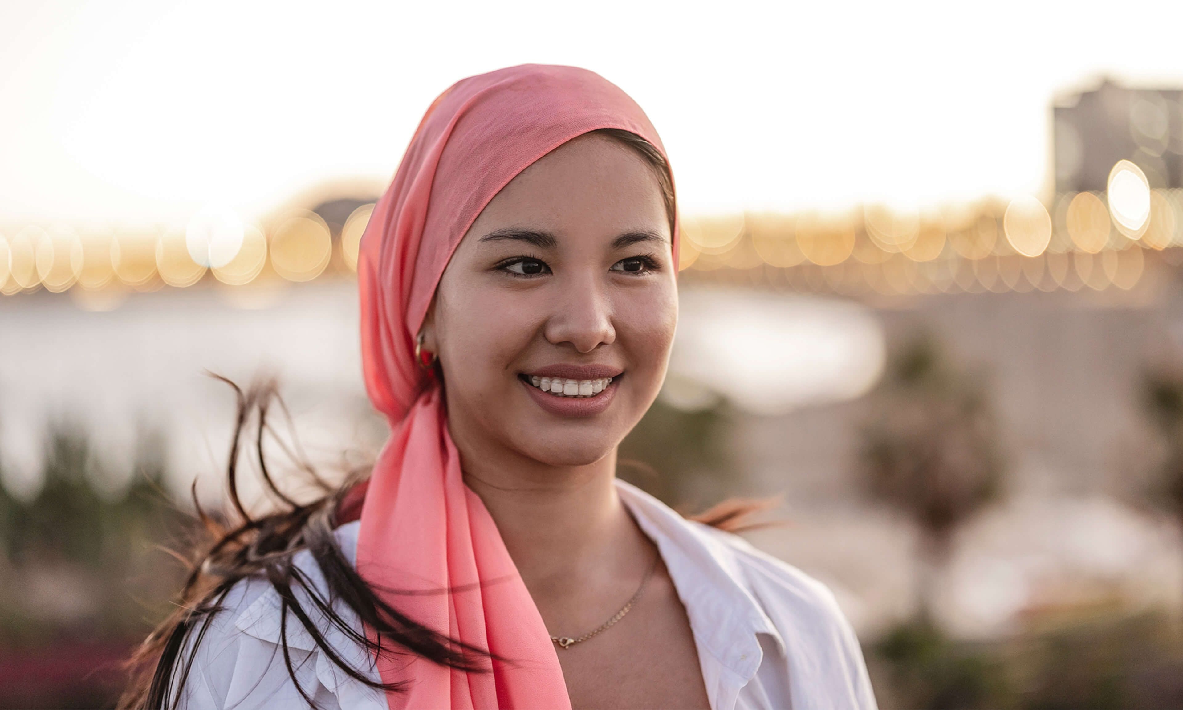 Una mujer sonriente con un pañuelo rosa en la cabeza y una blusa blanca de pie al aire libre durante el atardecer. El fondo incluye luces borrosas y vegetación, lo que crea una atmósfera serena.
