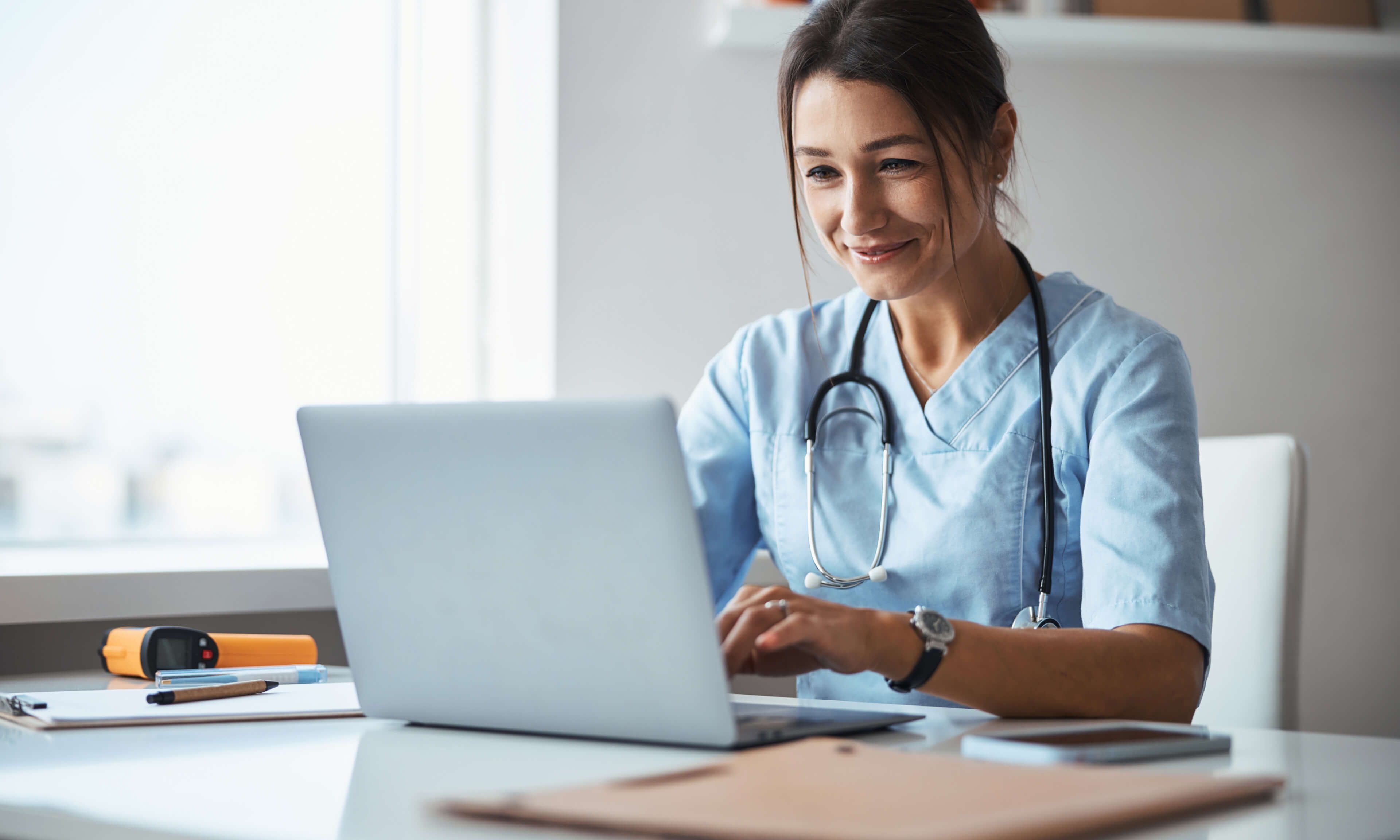 A nurse in scrubs works on a laptop at a desk in a brightly lit room.