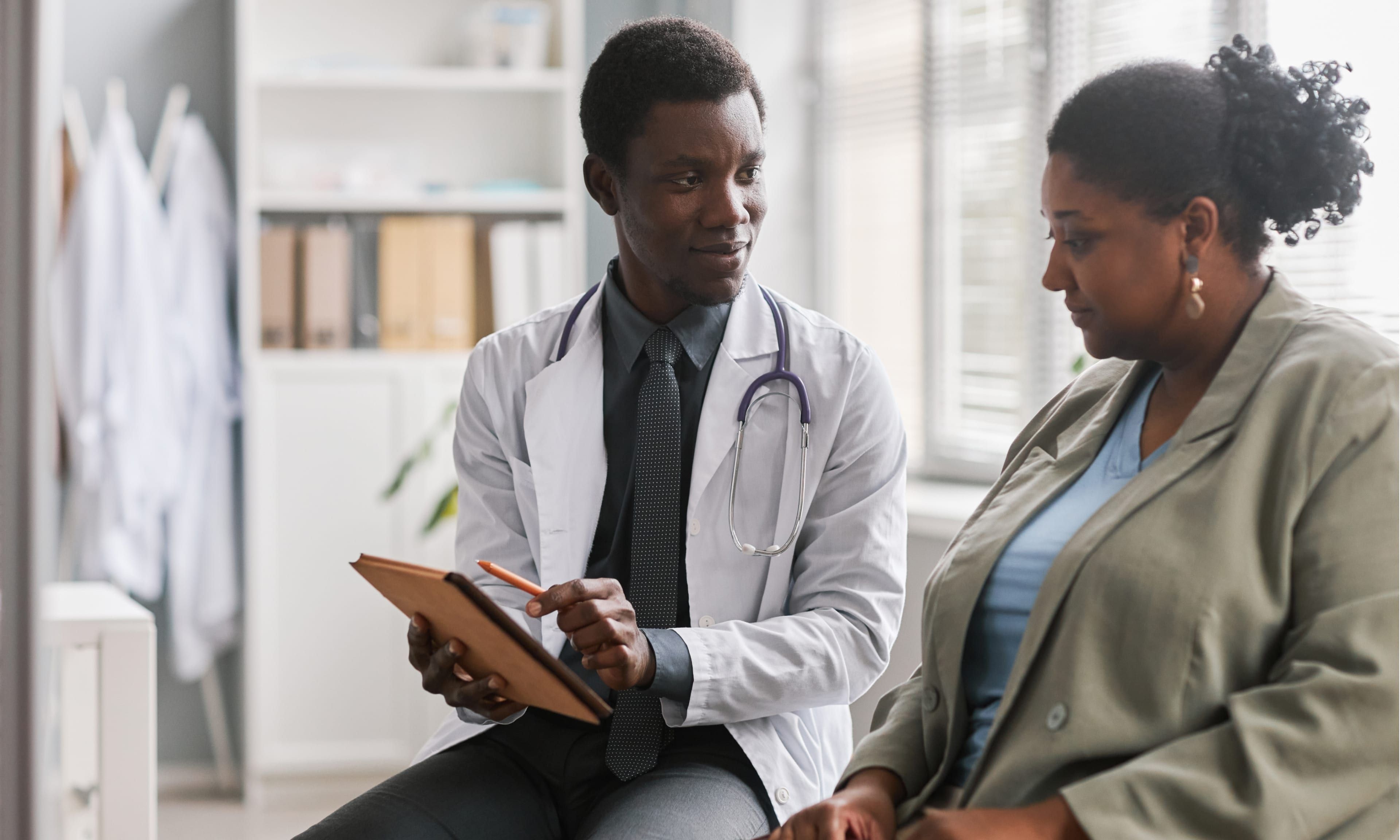 A doctor with a stethoscope consults a patient, holding a tablet in a medical office.