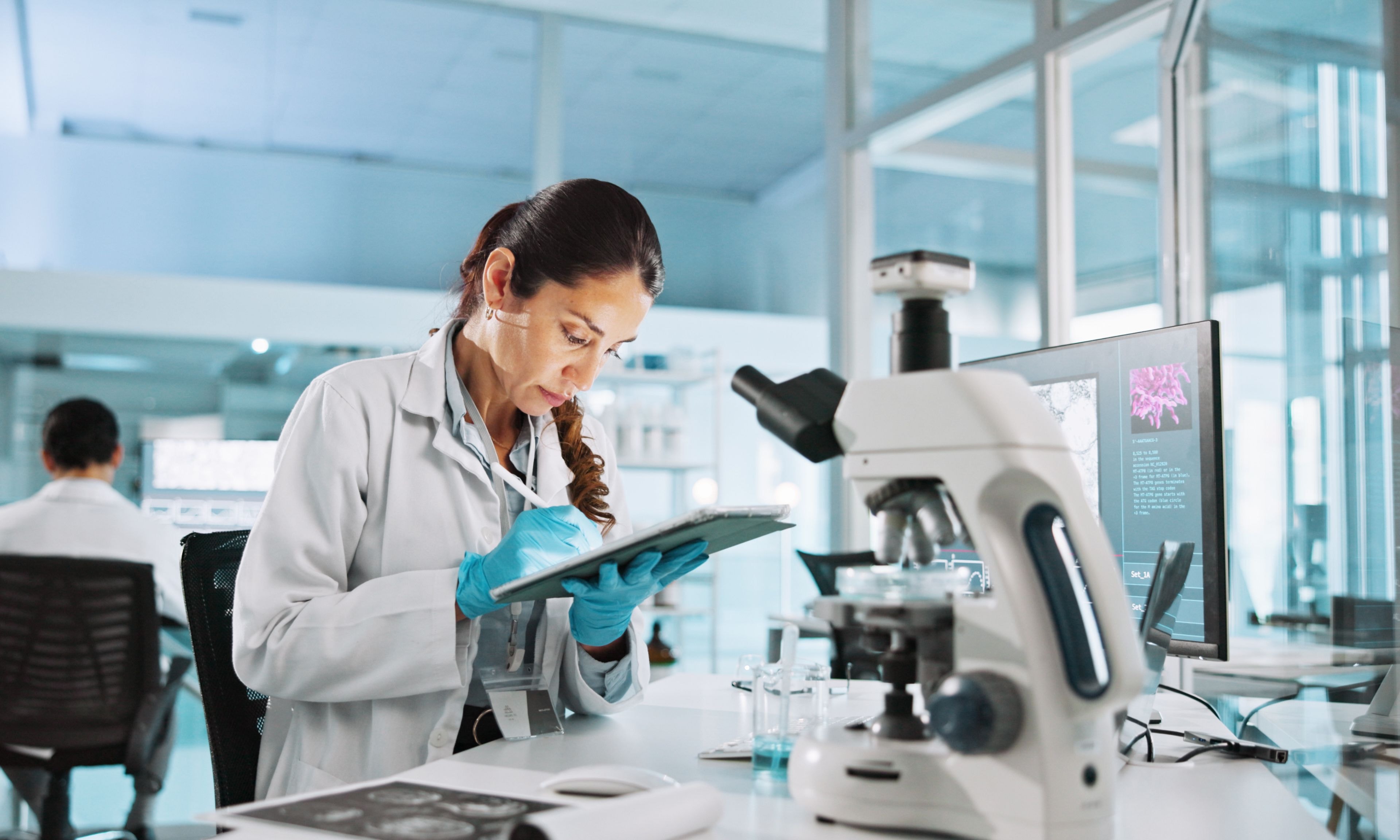 A scientist in a lab takes notes on a tablet next to a digital microscope.