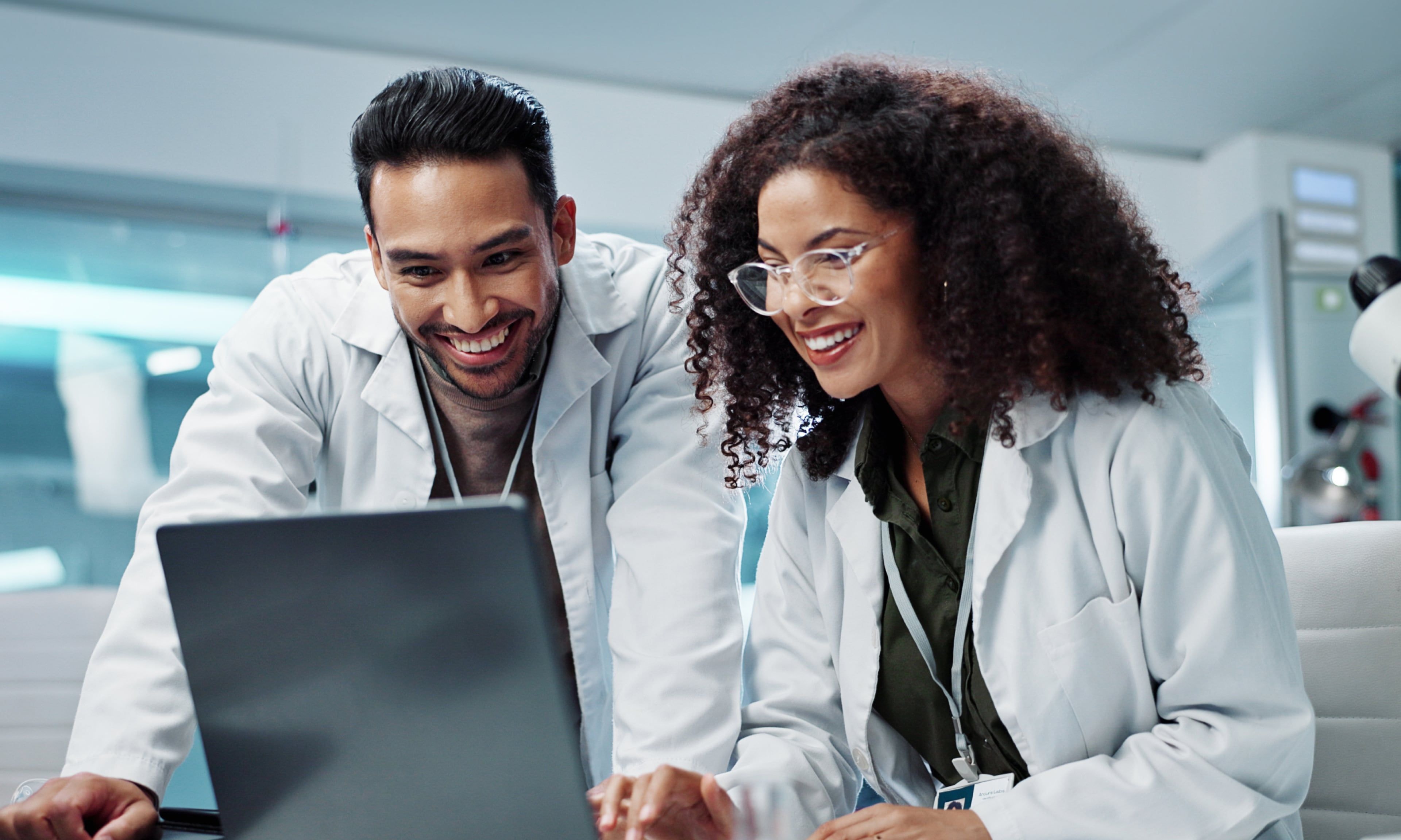 Two scientists in lab coats analyze data together on a laptop inside a modern laboratory.