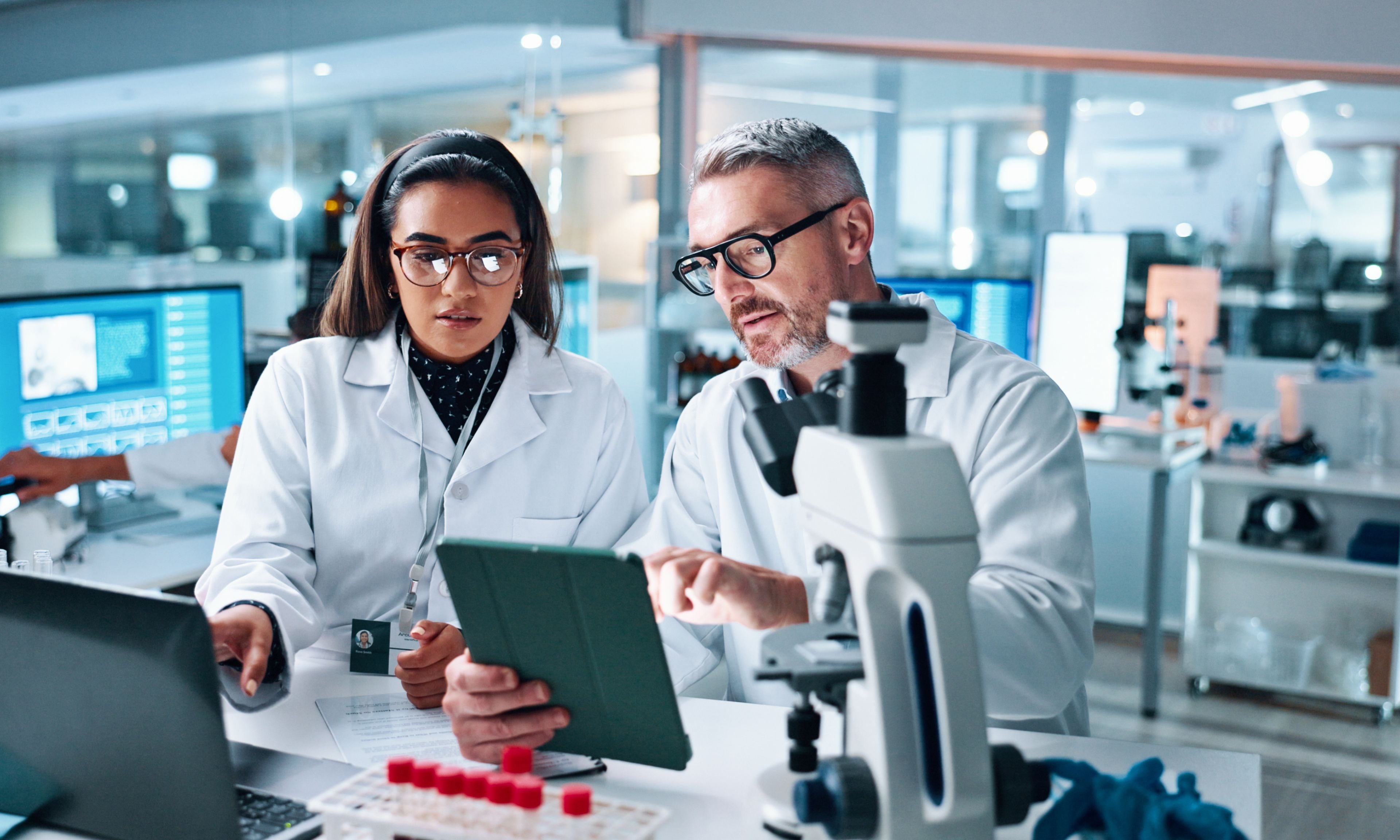 Two researchers in a lab review data on a tablet beside a microscope.