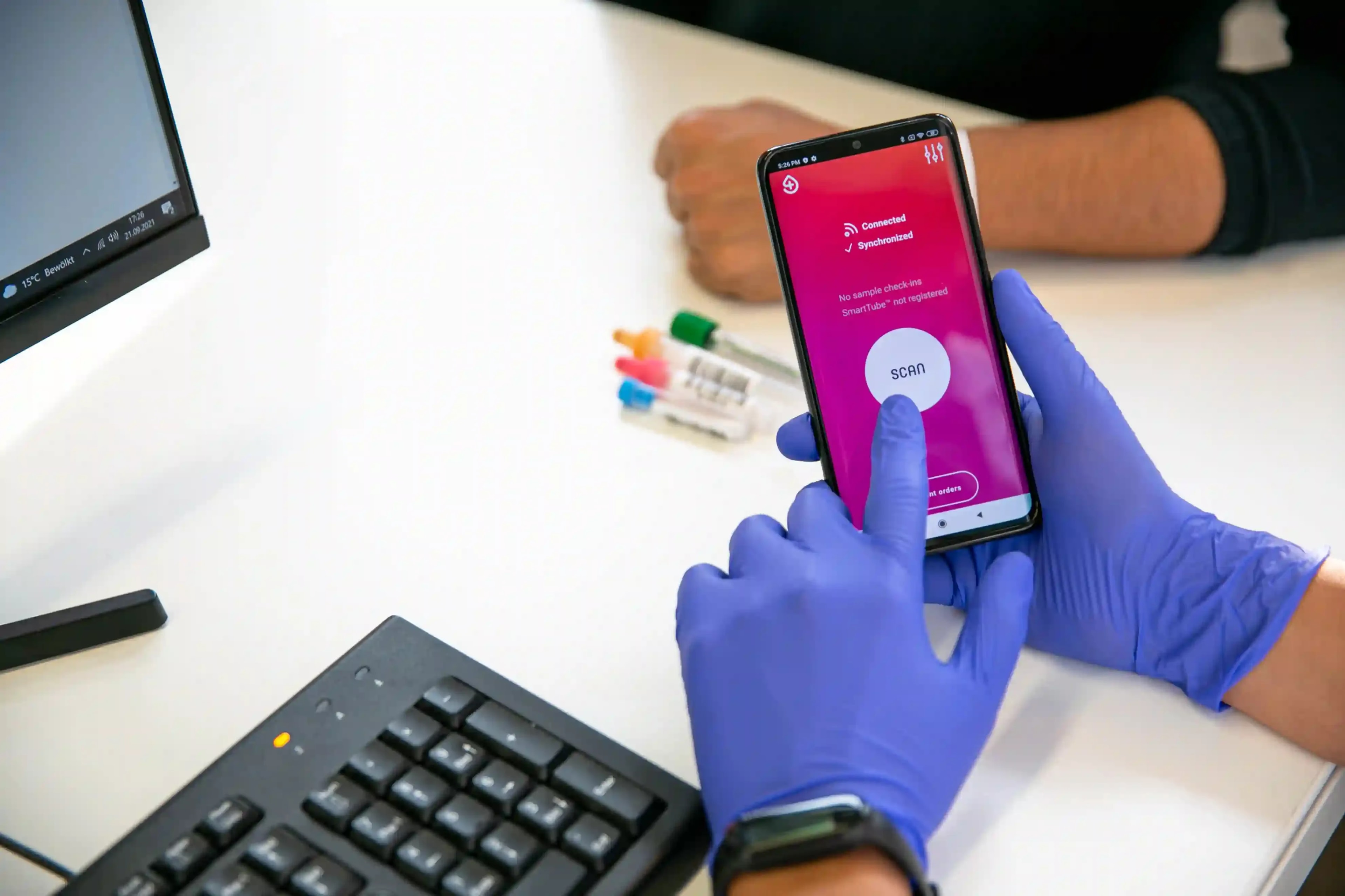 A healthcare professional wearing blue gloves uses the Smart4Diagnostics app on a smartphone to scan and monitor patient samples, reducing pre-analytical errors.