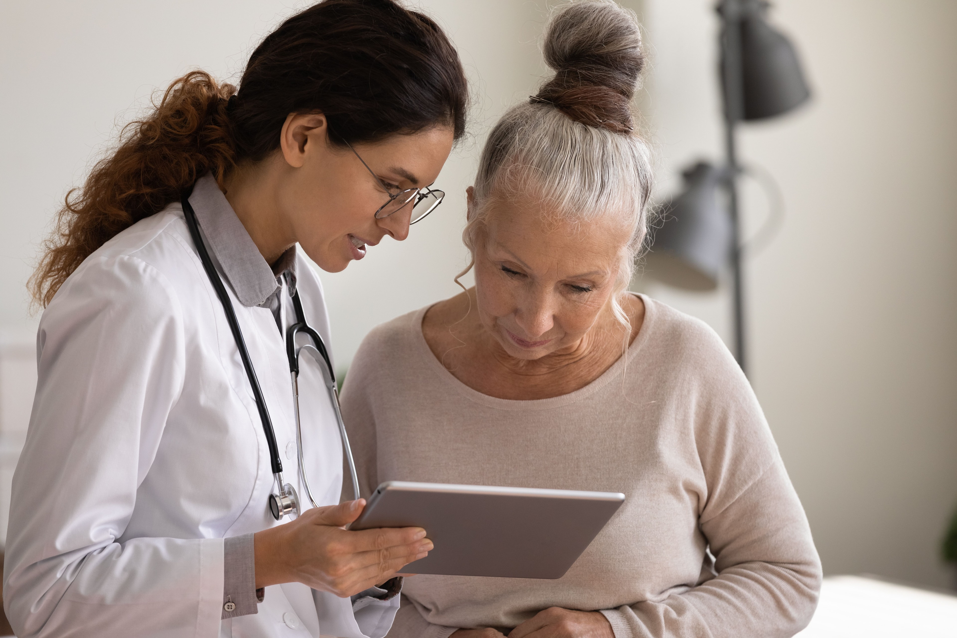 Medizinische Fachkraft, die mit einer älteren Patientin während eines Gespräches Informationen auf einem Tablet durchgeht.